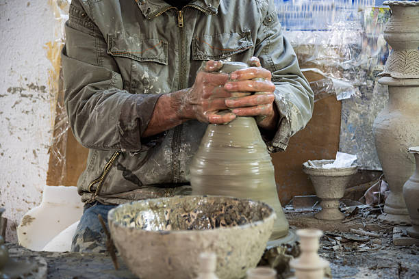 A Moroccan potter shapes clay on a dusty table in a Fes workshop, surrounded by finished and in-progress pieces. The scene radiates craftsman focus and traditional pottery artistry in Morocco.
