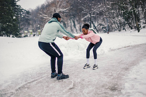 African american couple training on a snowy park path, man supporting woman as she practices ice skating squats, teamwork, balance and winter fitness outdoors
