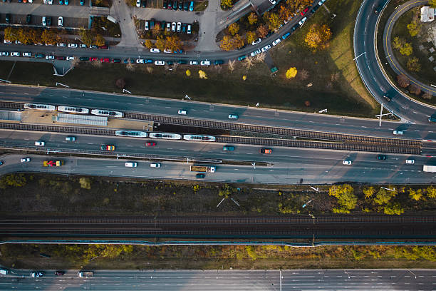 Top-down aerial image showing a multilane roadway with moving vehicles and tram lines running through the center, flanked by green areas and a railway track below. Curving roads, parking lots, and autumn-colored trees frame the urban transportation network, emphasizing the complexity and organization of city traffic systems.