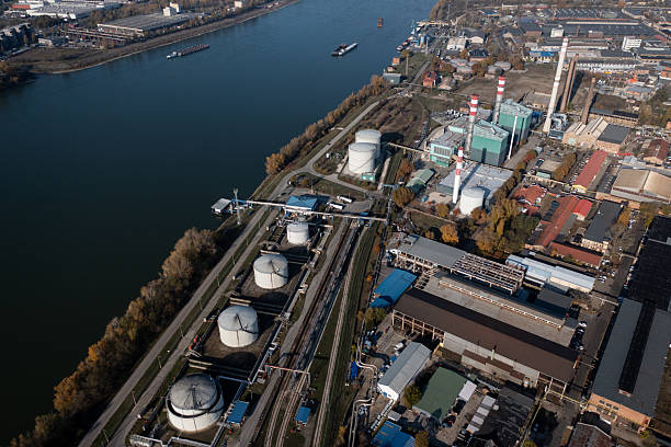 Aerial photograph of a large industrial complex featuring cylindrical storage tanks, pipelines, and factory buildings located along a riverbank. Multiple smokestacks and industrial infrastructure dominate the scene, while barges move along the calm river. The layout illustrates the integration of transportation and production facilities in a key logistical location.