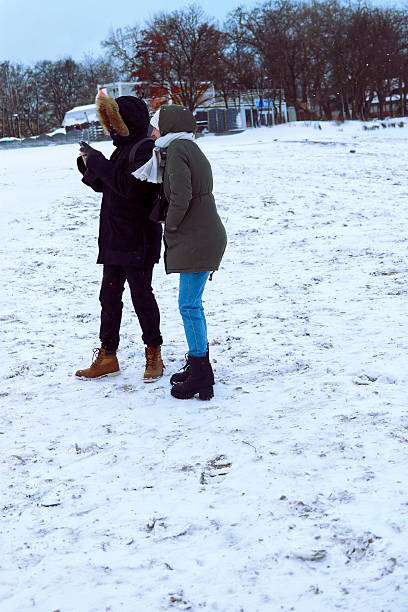Gdansk, Poland - January 11, 2026: Two people in winter coats standing on a snowy beach looking at a cell phone