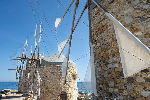 Old traditional windmills at beach of Chios Town on Chios Island in Greece on a on sunny day in late summer (or early autumn), with sea waves and light puffy clouds. Windmills have been used on Greek islands for grinding cereal grains for hundreds of years. These mills were originally built more than 100 years old and has been restored in recent years. For the mills to function, sails are mounted between the wooden spikes.