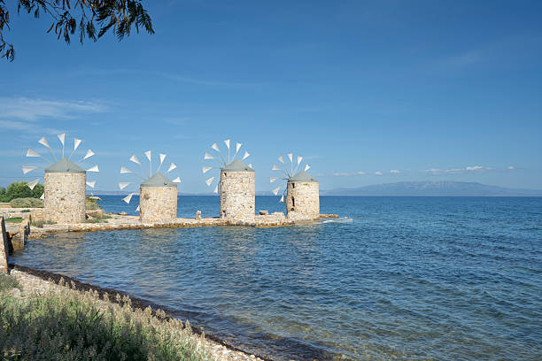 Old traditional windmills at beach of Chios Town on Chios Island in Greece on a on sunny day in late summer (or early autumn), with sea waves and light puffy clouds. Windmills have been used on Greek islands for grinding cereal grains for hundreds of years. These mills were originally built more than 100 years old and has been restored in recent years. For the mills to function, sails are mounted between the wooden spikes.