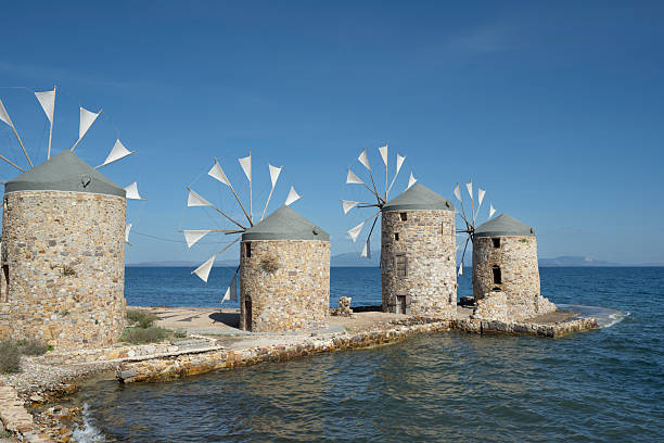 Old traditional windmills at beach of Chios Town on Chios Island in Greece on a on sunny day in late summer (or early autumn), with sea waves and light puffy clouds. Windmills have been used on Greek islands for grinding cereal grains for hundreds of years. These mills were originally built more than 100 years old and has been restored in recent years. For the mills to function, sails are mounted between the wooden spikes.