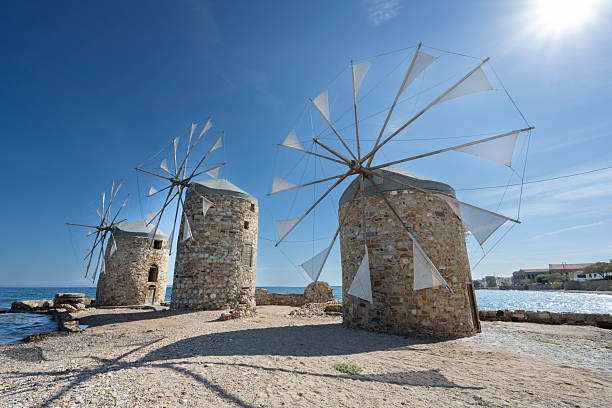 Old traditional windmills at beach of Chios Town on Chios Island in Greece on a on sunny day in late summer (or early autumn), with sea waves and light puffy clouds. Windmills have been used on Greek islands for grinding cereal grains for hundreds of years. These mills were originally built more than 100 years old and has been restored in recent years. For the mills to function, sails are mounted between the wooden spikes.