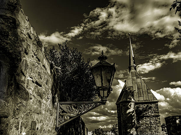 A contrasting black and white shot of the historic city wall. In the foreground on the left, the coarse texture of the quarry stones dominates, to which a cast iron lantern is attached with an artistic dragon mount. In the background, the dark, pointed silhouette of the Lukasklause rises under a dramatically cloudy sky.