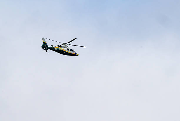 Newxcastle-upon-Tyne, UK: A yellow and white helicopter with green accents and rotor blades in motion soars through a clear blue sky. The aircraft features a streamlined fuselage, landing skids, and official markings suggesting emergency or police service. No people are visible in the image. The setting is outdoors in the daytime with bright natural light illuminating the scene. The focus is on the helicopter as the central subject against the expansive sky background, captured from a ground-level perspective. The image conveys aerial transportation or patrol activity in an open airspace environment.