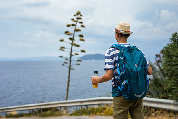 Teenage boy hiking along a coastal trail with sea view and a flowering agave tree in the background. Summer adventure, outdoor exploration, and scenic travel lifestyle.