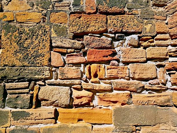 A rustic and unique stone wall featuring natural earth tones such as yellow, copper, orange, and sand, composed of stones in various sizes. The textured surface and warm colors create an organic, timeless background ideal for architectural, design, or natural concepts.