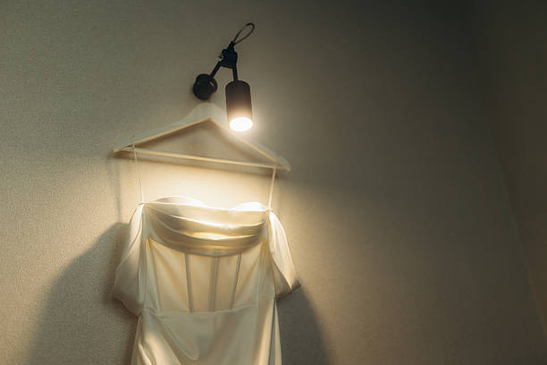 White bridal gown hanging on a hanger, illuminated by a warm spotlight on a light-colored wall, symbolizing preparation for marriage