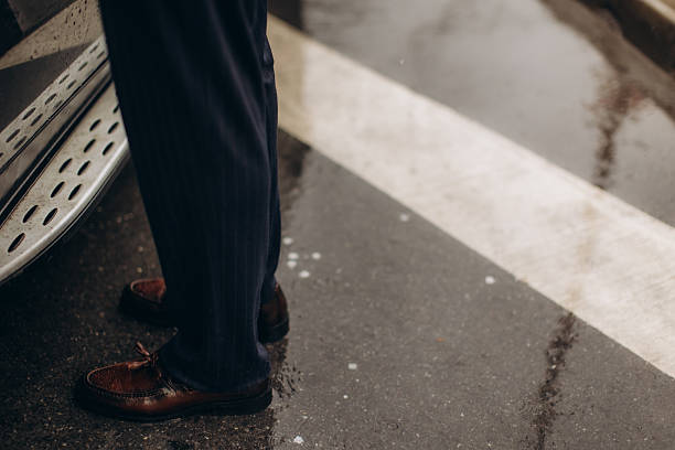 Man's legs with brown leather shoes and pinstripe trousers, standing in the rain on wet asphalt next to a car