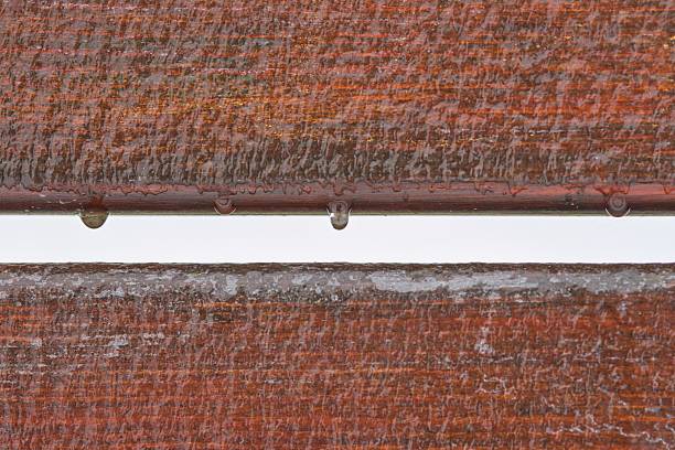 Close-up of water drops hanging from a wooden plank, showcasing the effects of rain or melting ice on a textured, red-brown surface.