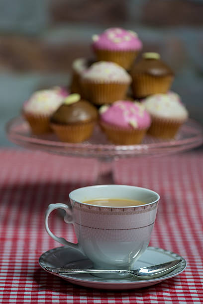 Cup of Tea and Cup Cakes on A Gingham Tablecloth. A white porcelain cup filled with tea rests on a matching saucer, accompanied by a silver spoon. The cup is placed on a red and white gingham tablecloth. In the background, a clear glass stand holds an assortment of cupcakes with various pastel-colored frostings, including pink and brown, decorated with small toppings. The setting suggests a cozy and inviting atmosphere, suitable for a casual tea time gathering.