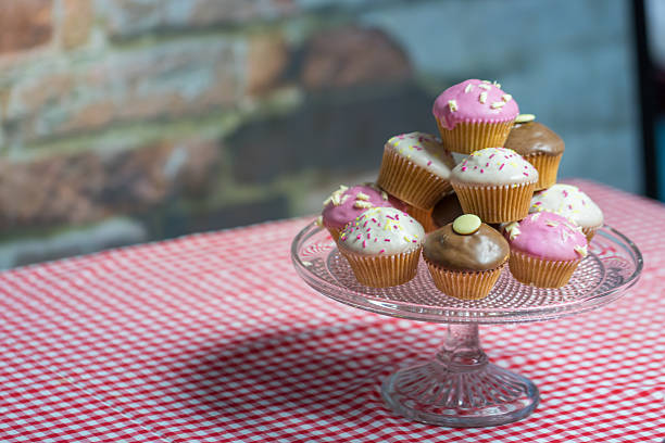Cup of Tea and Cup Cakes on A Gingham Tablecloth. A white porcelain cup filled with tea rests on a matching saucer, accompanied by a silver spoon. The cup is placed on a red and white gingham tablecloth. In the background, a clear glass stand holds an assortment of cupcakes with various pastel-colored frostings, including pink and brown, decorated with small toppings. The setting suggests a cozy and inviting atmosphere, suitable for a casual tea time gathering.
