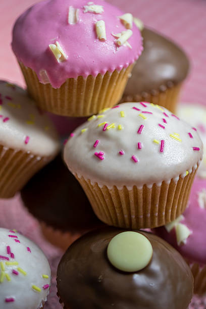 Cup of Tea and Cup Cakes on A Gingham Tablecloth. A white porcelain cup filled with tea rests on a matching saucer, accompanied by a silver spoon. The cup is placed on a red and white gingham tablecloth. In the background, a clear glass stand holds an assortment of cupcakes with various pastel-colored frostings, including pink and brown, decorated with small toppings. The setting suggests a cozy and inviting atmosphere, suitable for a casual tea time gathering.