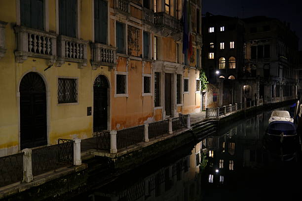 Night view of a quiet canal in Venice, Italy, lined with historic residential buildings. Warm light from windows reflects on the calm water, with small boats moored along the canal, creating a peaceful and atmospheric urban scene. Ideal for themes of travel, architecture, solitude, and European city life.