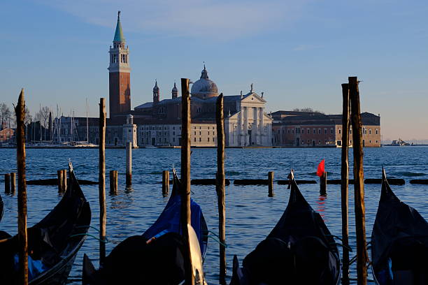 View across the Venetian lagoon with gondolas moored in the foreground and the island of San Giorgio Maggiore in the background, Venice, Italy. Calm water, wooden mooring poles, and historic architecture under a clear sky create a balanced and timeless travel scene. Ideal for concepts of tourism, culture, tradition, and European landmarks.