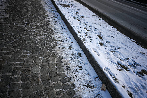 Wet cobblestone street next to a snow bank with footprints and an asphalt road