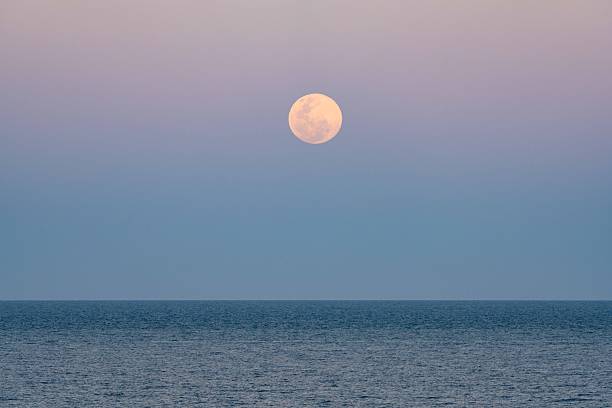 This striking coastal scene captures a bright December supermoon emerging over the ocean at sunset, with warm evening colours reflecting across the water and sky