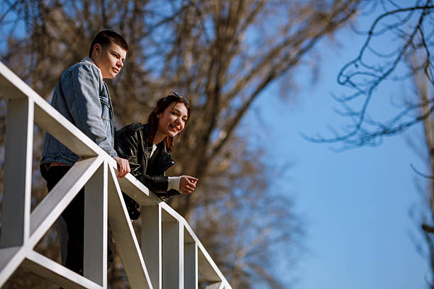 Two young adults are enjoying a sunny day at the park, leaning over a white railing. They are smiling and engaging in conversation during a relaxed afternoon in springtime.