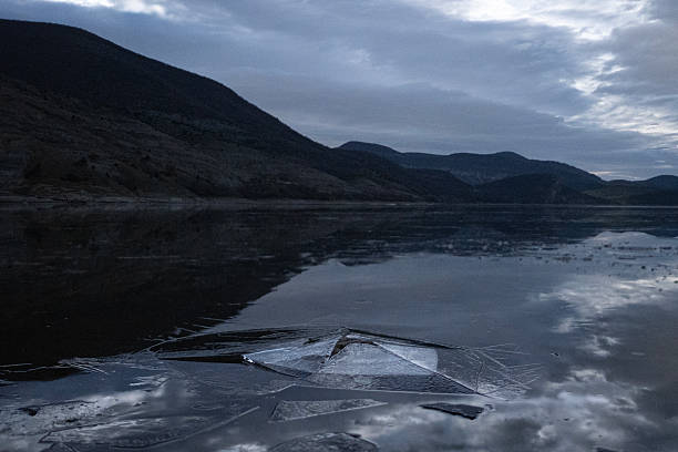Cracked ice structure melting on the surface of a partially frozen river, reflecting mountains and dramatic cloudy sky at twilight. night fantasy