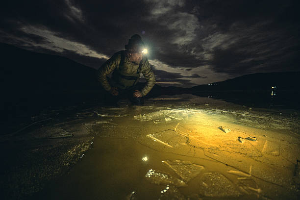 Mature fisherman with headlamp on extreme winter adventure exploring cracked ice on frozen water surface under dark night sky. Ice fishing in  mountain river