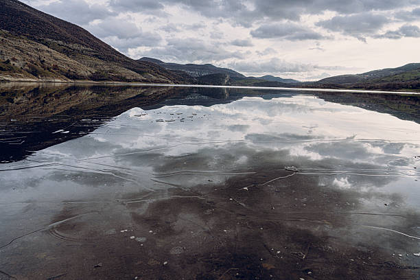 Frozen water surface reflecting morning sunlight and clouds creating surreal mirror. Nature background, mountain lake illuminated by soft sunlight