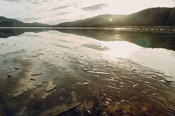 Frozen lake surface with ice shards and water reflecting mountains and morning sky under a bright sun, highlighting winter nature and climate