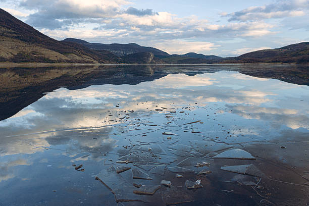 Still frozen water surface of a mountain lake with broken ice shards on the surface, reflecting the majestic sky and distant mountains in the morning. Landscape