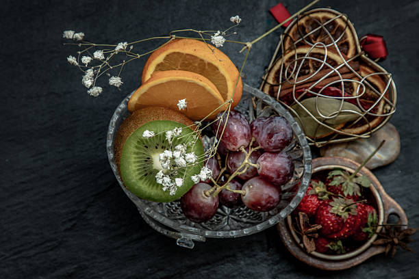 Artfully arranged on a dark background, a collection of fresh fruits like kiwi, oranges, grapes and strawberries mingles with dried citrus in different vessels.