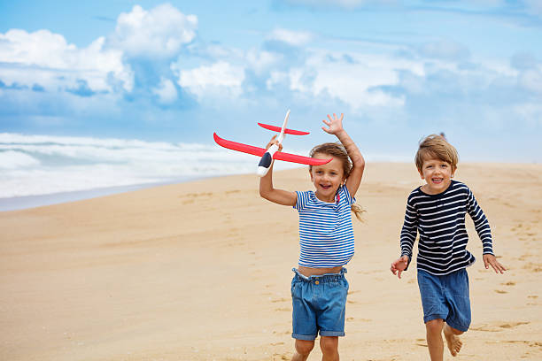 Two children boy girl, run excitedly along a sandy beach, one holding a toy airplane while the other smiles, with waves and clouds creating a cheerful atmosphere in the background summer vacation fun