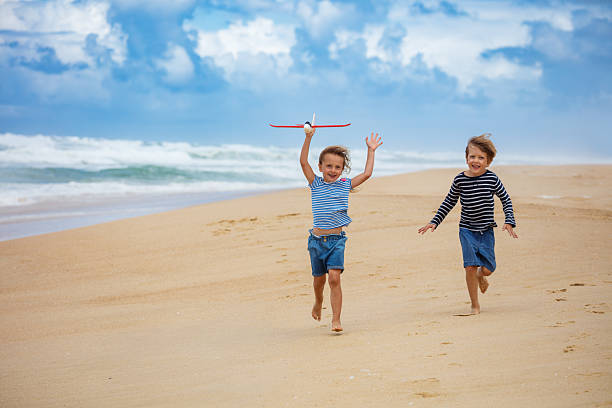Two children have vacations run along a sandy beach, one holding a toy plane while the ocean waves crash in the background under a beautiful cloudy sky