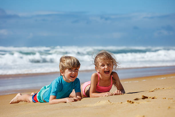 Two children are lying on the sandy beach on summer vacation, laughing joyfully while enjoying a sunny day by the ocean with waves crashing in the background and a clear blue sky above them