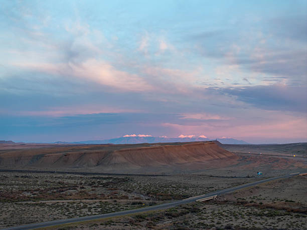 A breathtaking high-angle aerial view of a dramatic storm sunset in Green River, Utah. The image captures golden sunlight breaking through dark, moody clouds, creating visible rain shafts (virga) over the desert landscape. The iconic Book Cliffs mesa stands prominently on the right, while the small town and Interstate 70 are nestled in the valley below. A perfect representation of the wild, unpredictable weather of the American Southwest.
