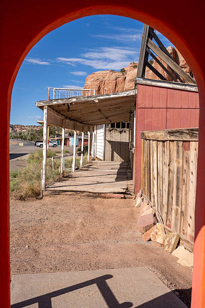 A weathered, abandoned roadside store under towering red sandstone cliffs. The dilapidated building features faded signs for silver jewelry and blankets, evoking a sense of nostalgia for mid-century American road travel and the historic Route 66 era. The contrast between the rugged natural mountains and the decaying man-made structure tells a story of the changing West