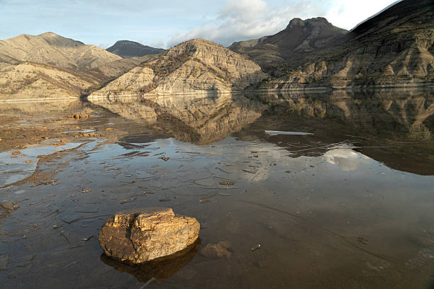 Still frozen water surface of a mountain lake with broken ice shards on the surface, reflecting the majestic morning sunlight, sky and distant mountains