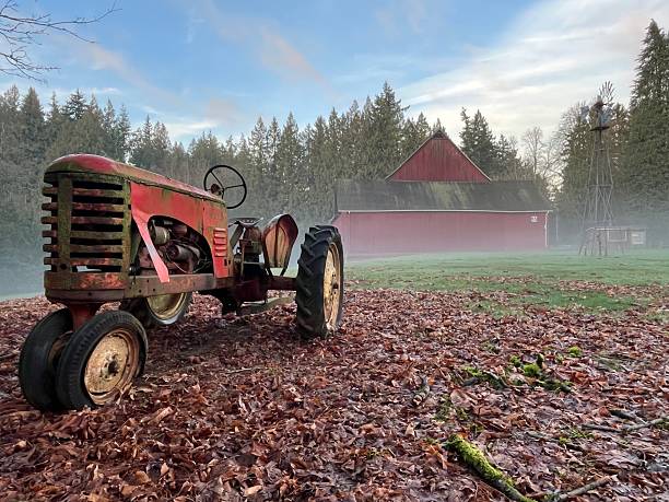 An old tractor sits on a ground covered with fallen leaves. A red barn stands in the background. Fog covers the area creating a misty scene. Trees surround the farm.