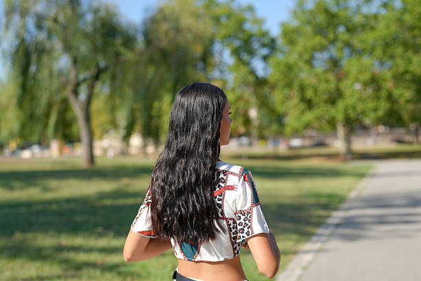 A woman with long hair is walking down a path in a park. The park is filled with trees and grass, and there are cars parked in the background. The woman is wearing a white shirt and blue pants