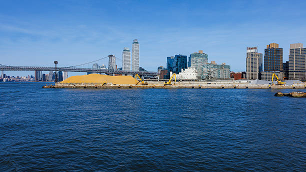Industrial waterfront on the East River in New York City with piles of sand and gravel for construction. The Williamsburg Bridge and the Brooklyn skyline are visible in the background on a sunny day.