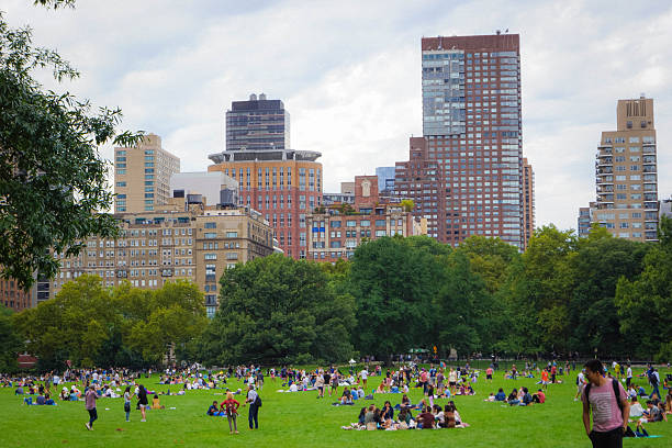 Central Park Great Lawn with crowds picnicking amid trees and Upper West Side skyline on summer day at New York City, USA.