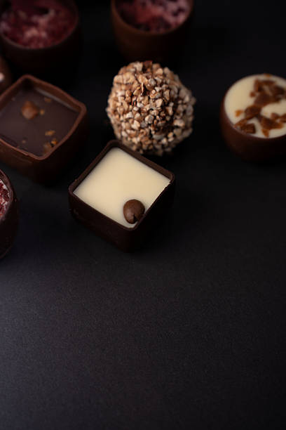 close-up of artisanal chocolates on a matte black surface, featuring a white-cream square topped by a coffee bean, a nut-crusted truffle and glossy dark pralines in moody lighting, framing