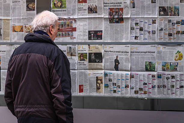 Ljubljana, Slovenia December 27, 2025, A person is standing in front of a glass display covered with multiple pages of newspapers. The newspapers are arranged in rows and columns, showcasing various articles, images, and advertisements. The person is wearing a dark jacket and has light-colored hair, viewed from behind. The setting appears to be an urban area, possibly near a newsstand or public information board.