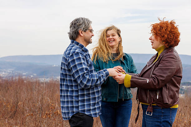 A mature heterosexual couple, a 46-year-old redhead woman and a 54-year-old man, hold hands during their informal outdoor wedding ceremony. A 50-year-old female minister officiates the service. The bride and minister are talking while the groom smiles. The ceremony takes place in the Poconos mountains of Pennsylvania during a cloudy afternoon in early spring. The background shows rolling hills and dormant vegetation. Medium shot.