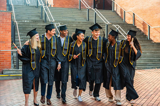 A large group of university graduates huddle in closely together outside as they pose for a portrait.  They are dressed in formal black gowns and have caps on as they hold their diplomas.