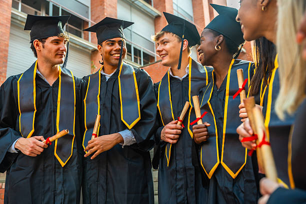 Group of diverse university students wearing graduation caps and gowns, holding diplomas, smiling and interacting on their graduation day at the campus