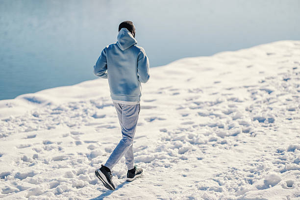 Adult man jogging in winter sportswear along a snow-covered lakeside path, back view showing footprints in fresh snow, enjoying outdoor fitness and cold-weather training