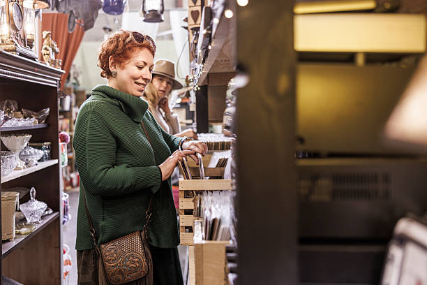 A 46-year-old redhead woman smiles while browsing for items in a crowded antique store. Her 51-year-old friend, wearing a hat, shops in the background. The shot has selective focus on the woman in the foreground, with shelves of vintage goods visible.