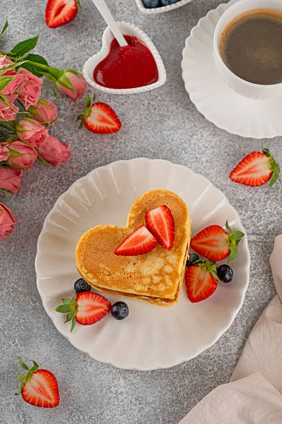 Heart-shaped pancakes with fresh strawberries and blueberries on elegant plate, served with coffee, pink roses and berry sauce. Valentine's Day breakfast or brunch. Romantic food concept. Top view, copy space