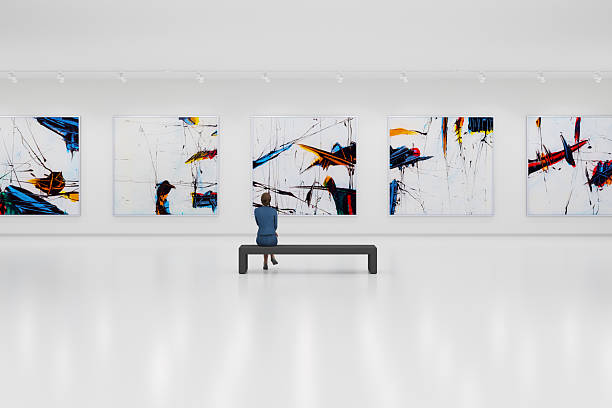 The interior of a modern and minimalist art gallery. The floors and walls are white, with large abstract expressionist paintings in black, blue, yellow, and red hanging on the white walls. A  visitor looking at the paintings.