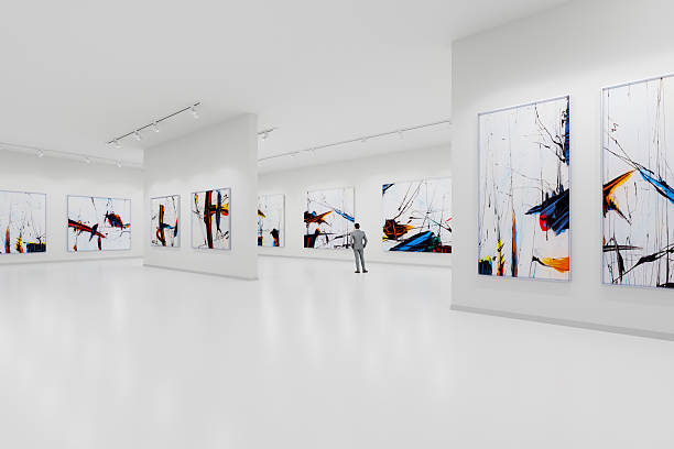 The interior of a modern and minimalist art gallery. The floors and walls are white, with large abstract expressionist paintings in black, blue, yellow, and red hanging on the white walls. A man is looking at the paintings.